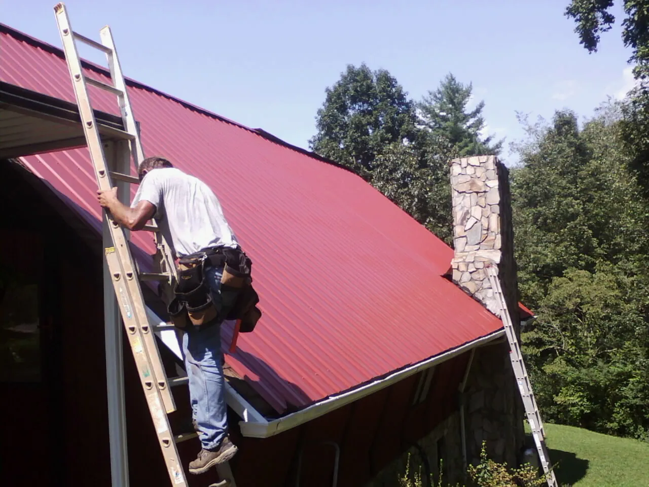 Roofer on a ladder beside a red metal roof and stone chimney — Soffit & Fascia Repair by Roofing Brothers in Kaplan